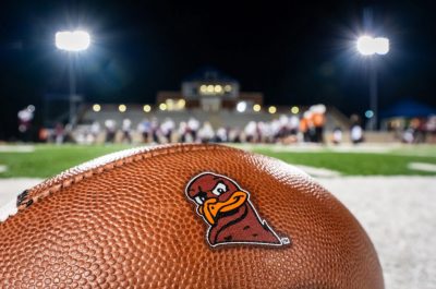 The logo of the HokieBird appears on a football in the foreground under football lights.